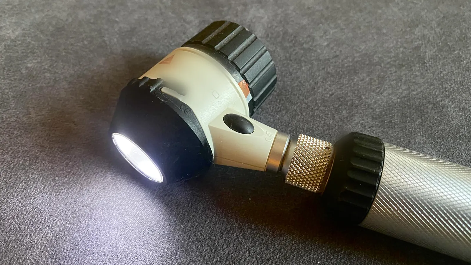 Dermatoscópio de mão com a luz acesa, apoiado sobre uma superfície cinza, sem pessoas na imagem.